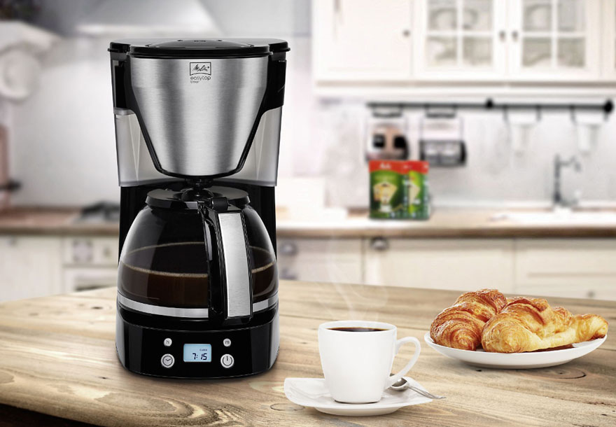 Een zilveren koffiezetapparaat naast een wit kopje koffie en croissants op een houten tafel in een keuken.