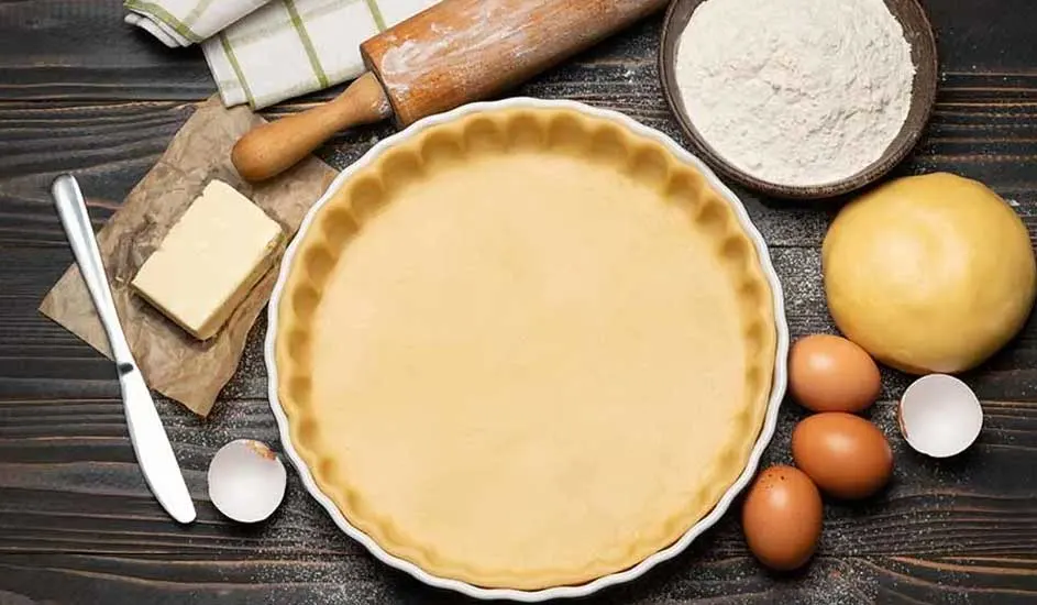 Een taartbodem in een witte schaal, met bloem, boter, eieren, deeg en een rolstok op een houten tafel.