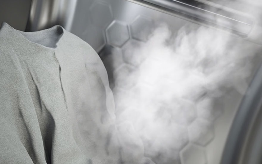 Un vêtement gris clair est suspendu dans un appareil métallique avec des motifs hexagonaux. De la vapeur s'en échappe.