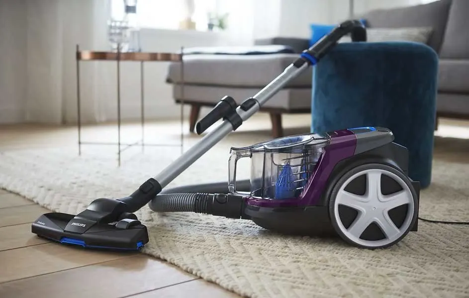 Un aspirateur traîneau violet et noir sur un tapis à motifs dans un intérieur lumineux.