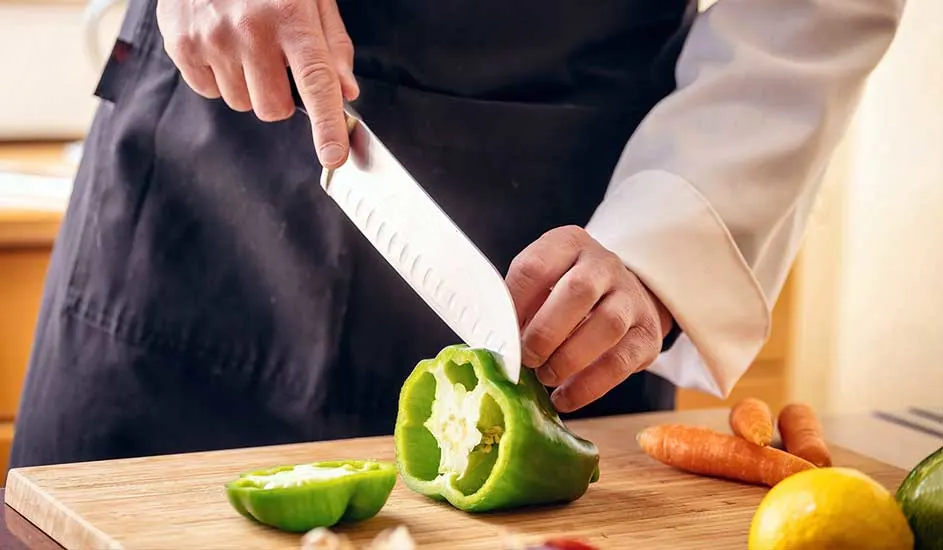 Un cuisinier en tablier coupe un poivron vert sur une planche à découper en bois. Des carottes et un citron sont à proximité.