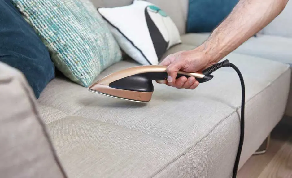 Hand strijkt een beige sofa met een goudkleurig strijkijzer. Kussens in blauw en wit liggen op de achtergrond.