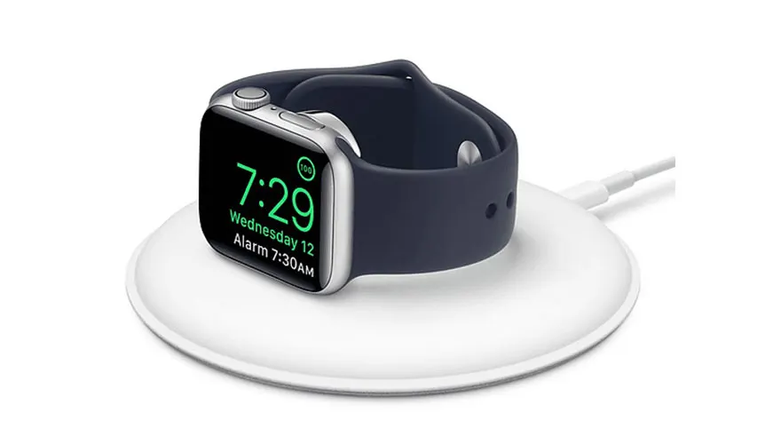 Een zilverkleurige smartwatch met een blauw bandje ligt op een witte draadloze oplader. Het scherm toont de tijd 7:29.