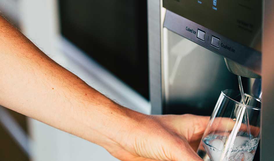 Une main tient un verre sous le distributeur d’eau d’un réfrigérateur en inox, tandis qu’un filet d’eau fraîche le remplit.