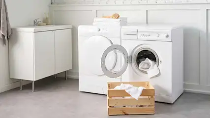 Witte wasmachine en droogkast in een wasruimte. Een houten bak met wasgoed staat op de grijze vloer.
