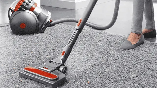 Aspirateur gris et rouge sur un tapis gris à poils longs. Un tuyau relie l'aspirateur à la tête de nettoyage.