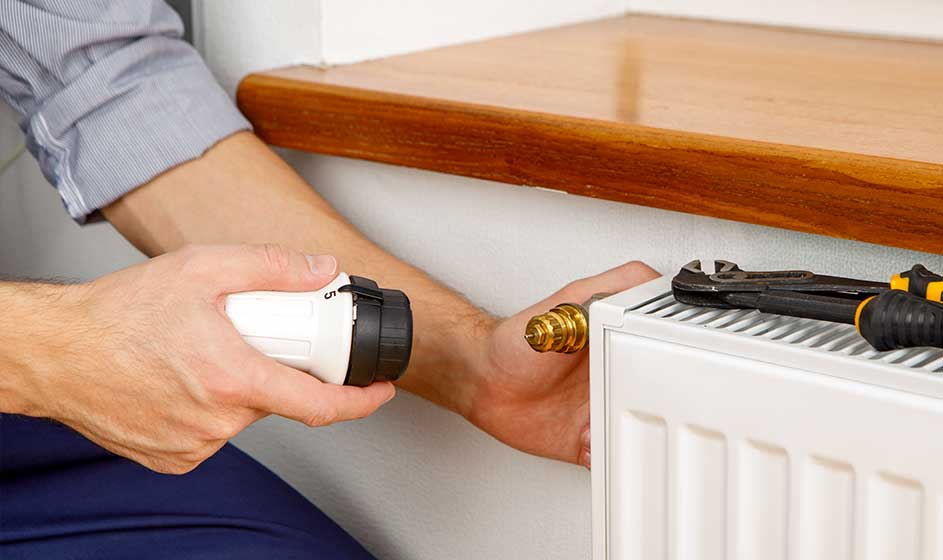 Une personne dévisse une vanne blanche de radiateur tandis qu’une clé à molette repose dessus. Le système de chauffage est en cours d’entretien.
