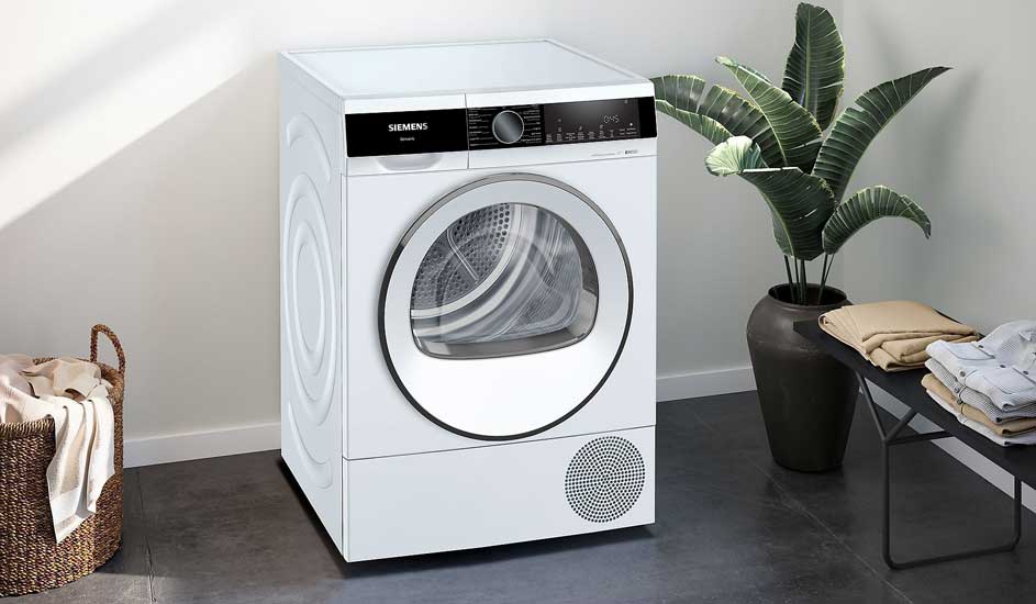 Un sèche-linge blanc moderne de Siemens se trouve dans une pièce ordonnée, à côté d'une plante, d'un panier à linge et de vêtements pliés.