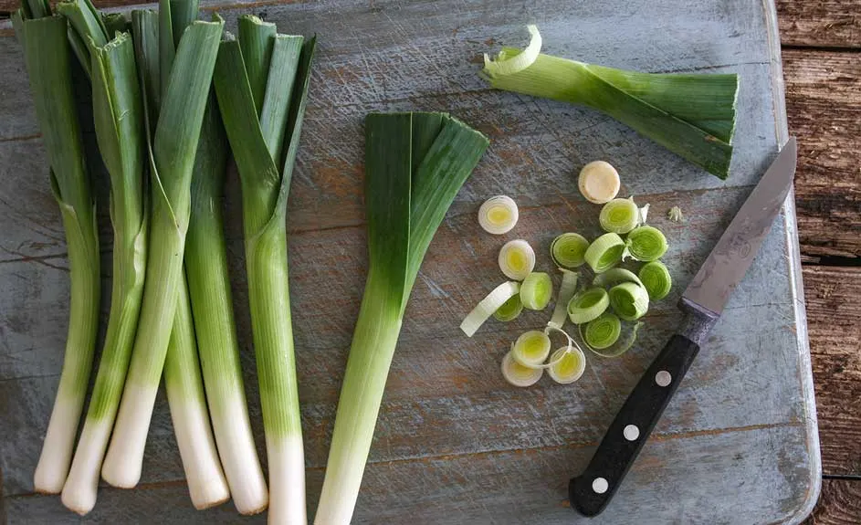 Des poireaux verts et blancs entiers et coupés sur une planche à découper en bois bleue avec un couteau.