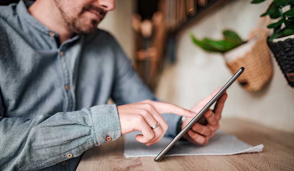 A man in a light blue shirt types on a tablet at a wooden table, with plants and cosy decorations in the background.