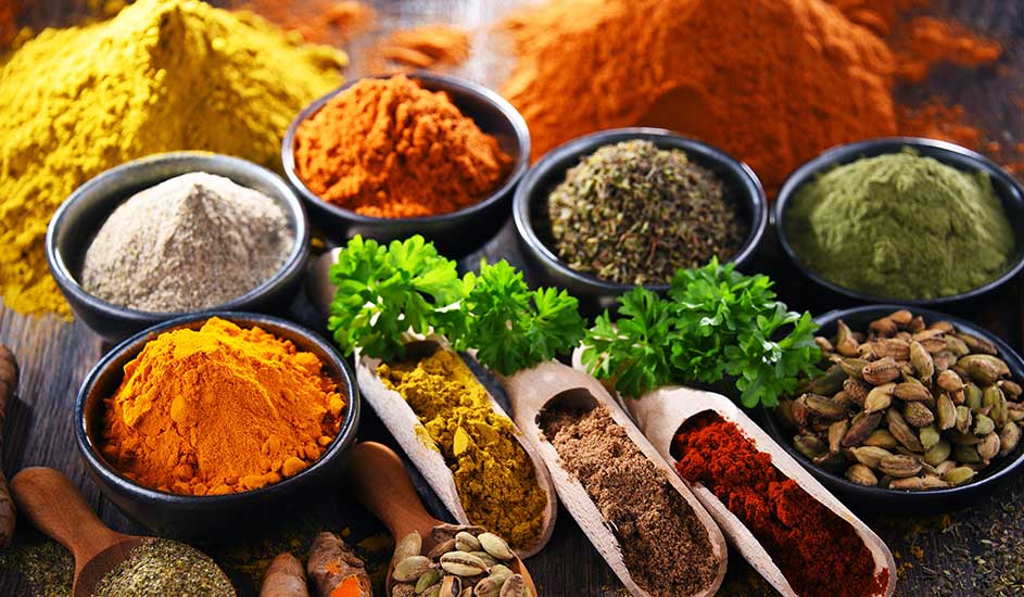 Diverse kruiden en specerijen in kommen en houten lepels, met levendige kleuren zoals geel, oranje, groen en bruin.