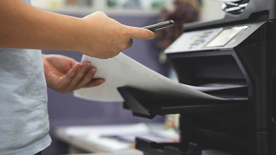 Een persoon legt wit papier in een zwarte printer. De achtergrond is vaag en lichtpaars.