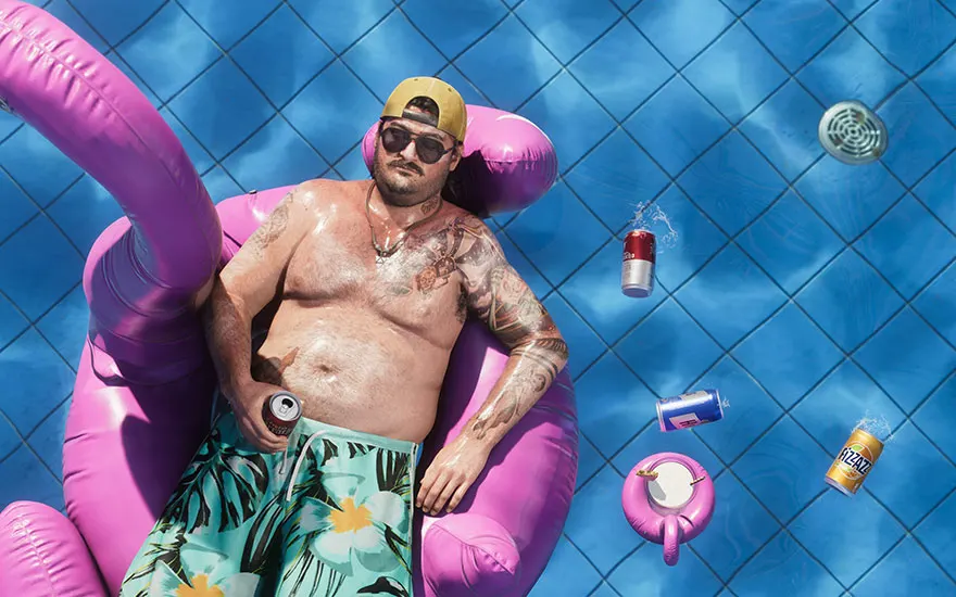 Man relaxing on a pink inflatable flamingo in a swimming pool with drink cans around him.