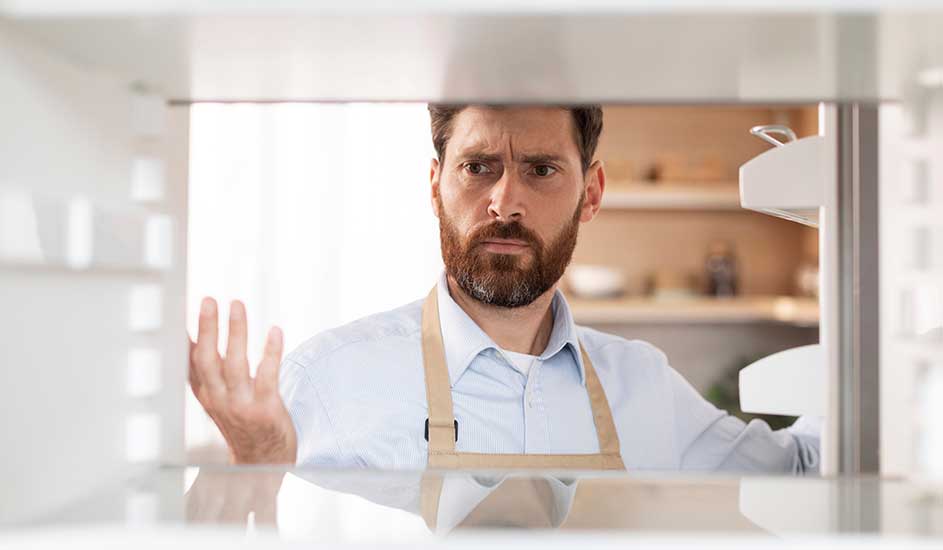 Een man met een baard, gekleed in een schort, staat voor een open koelkast in een keuken. De schappen zijn grotendeels leeg.