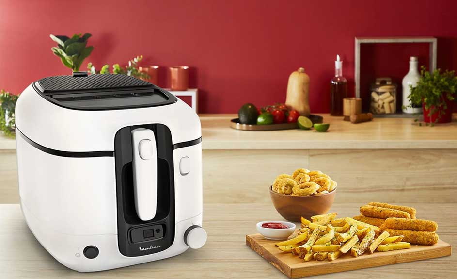 Een witte friteuse staat naast een houten plank met friet en snacks, tegen een rode achtergrond.