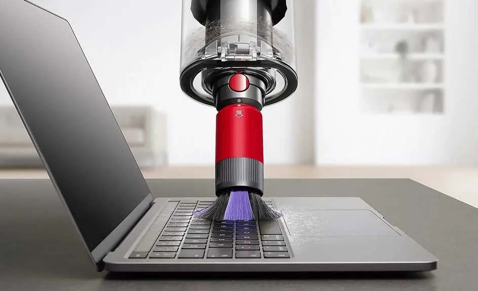 Een grijze laptop wordt schoongemaakt met een rode stofzuiger op een grijs oppervlak. Een witte kast is vaag op de achtergrond te zien.
