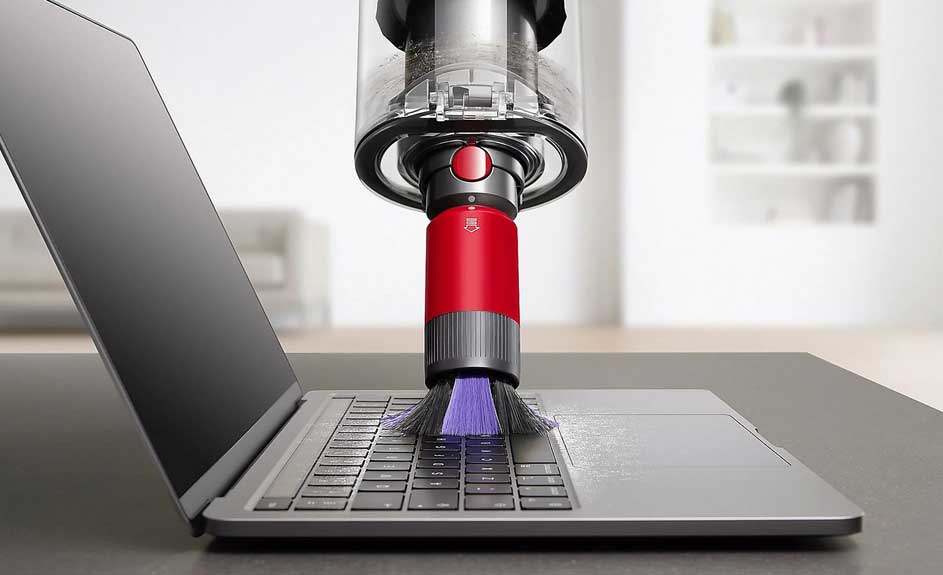 Un ordinateur portable gris est nettoyé avec un aspirateur rouge et transparent sur une table grise.