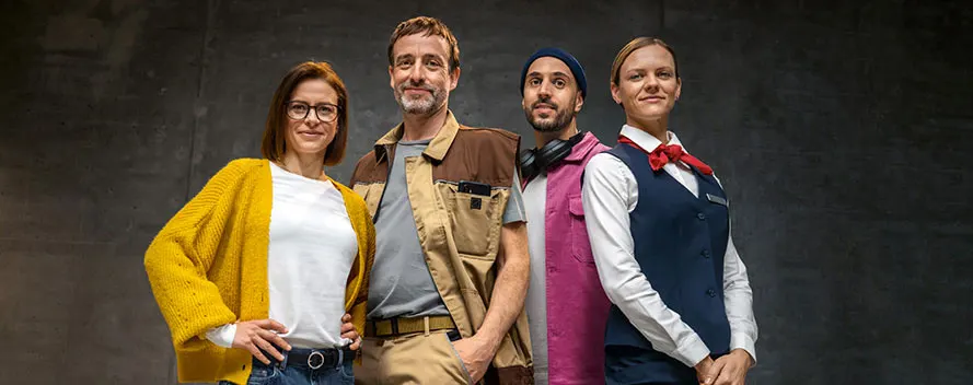 Four people in work clothing stand against a dark gray background. The clothing includes a yellow cardigan, brown work jacket, pink shirt, and navy vest.