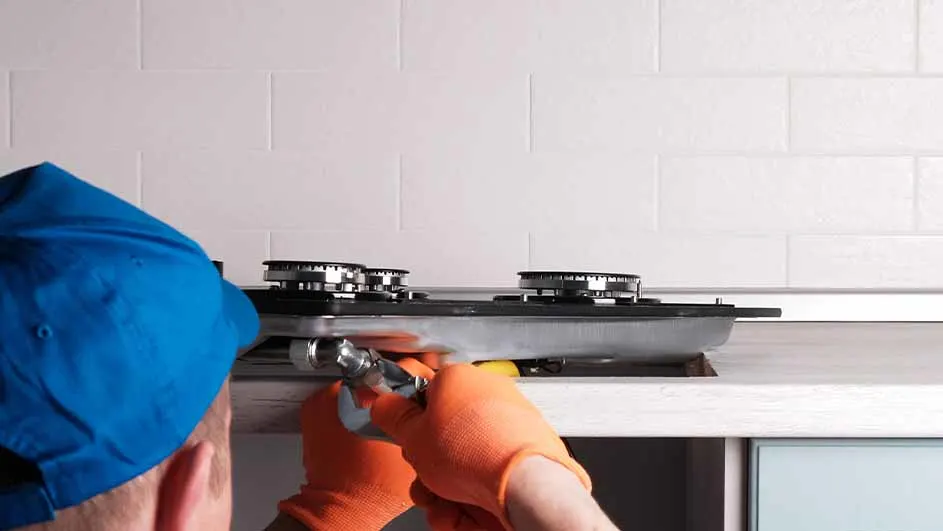 Een persoon met oranje handschoenen repareert een gasfornuis met een sleutel tegen een witte tegelachtergrond.