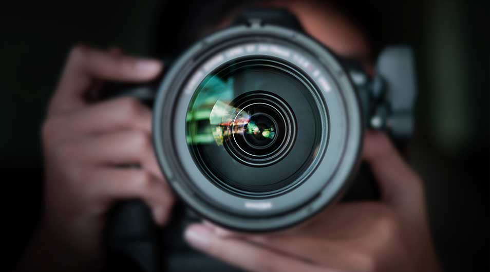Een close-up van een zwarte cameralens, met reflecties zichtbaar op het glas. Handen houden de camera vast tegen een donkere achtergrond.