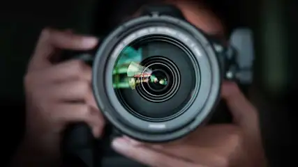 Un gros objectif d'appareil photo noir au premier plan, tenu par des mains floues, sur un fond sombre.