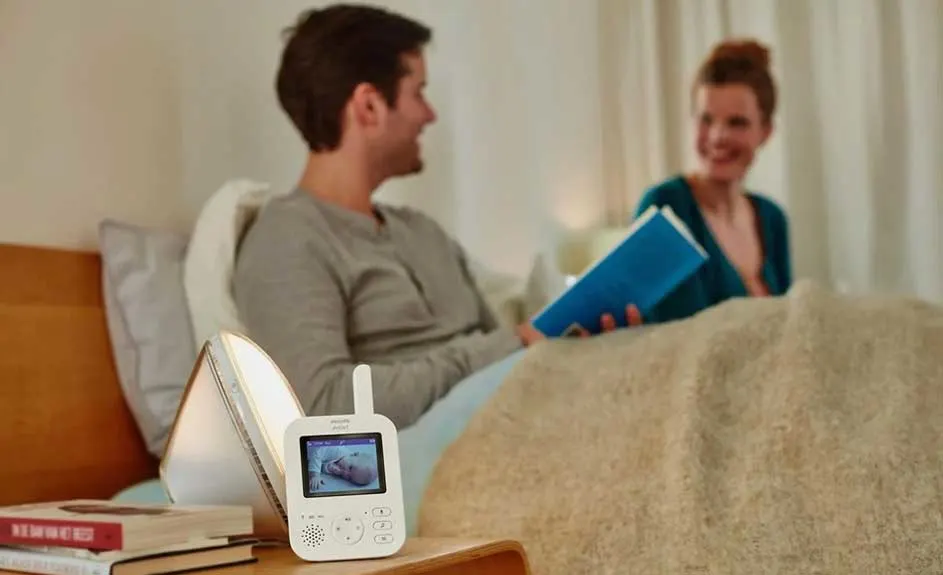 Un babyphone blanc sur une table de chevet en bois, près d'une lampe et de livres, dans une chambre à coucher avec un lit.