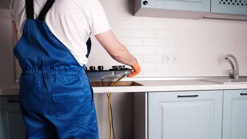 A kitchen scene shows blue cabinets, a white countertop, and a cooktop being installed. A yellow wire hangs below the counter.