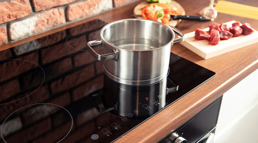 Een zilveren kookpot met water staat op een zwarte inductiekookplaat naast een houten snijplank met vlees.