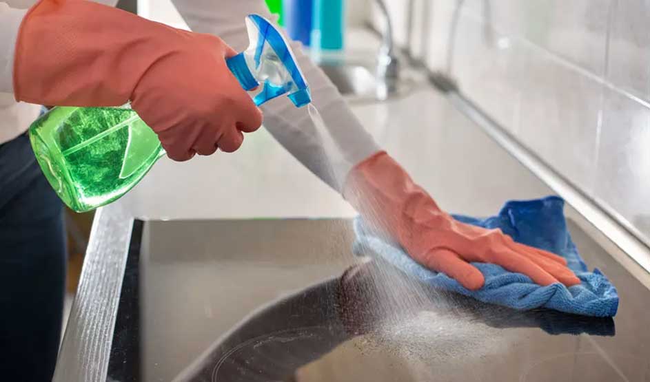 Une personne nettoie une plaque de cuisson noire avec un chiffon bleu et un vaporisateur vert. Des gants protègent ses mains.