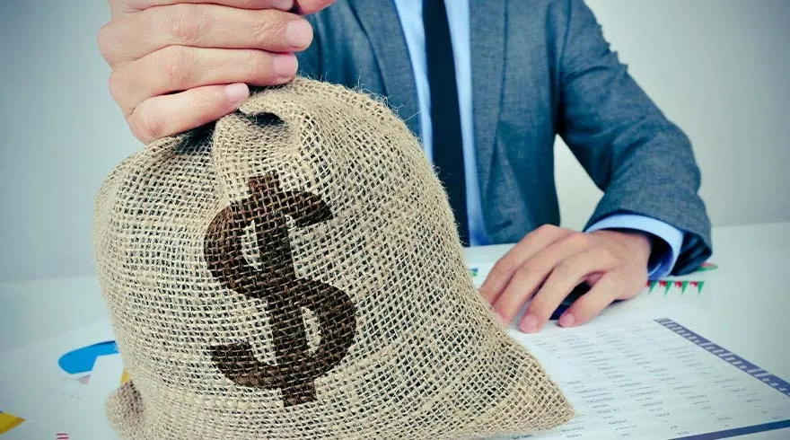 Sac en toile de jute avec un symbole dollar brun, sur une table blanche avec des documents et un homme en costume en arrière-plan.