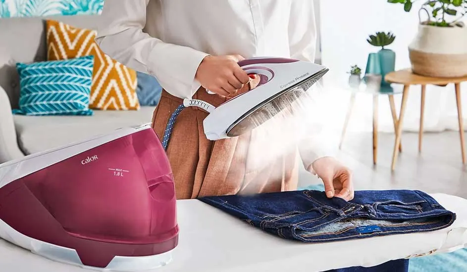 Een stoomstrijkijzer strijkt een blauwe jeans op een witte strijkplank, met een Calor-stoomgenerator op de achtergrond.
