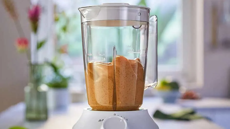 Un mixeur blanc avec une carafe transparente contenant un mélange beige est sur une table, devant une fenêtre lumineuse.