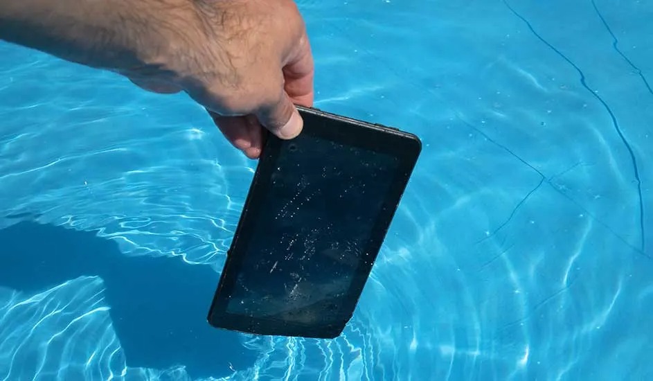 Een hand houdt een zwarte tablet net boven het wateroppervlak van een zwembad, merk is niet zichtbaar.