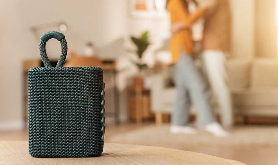 Op de voorgrond staat een compacte, gevlochten zwarte luidspreker met draaglus, terwijl iemand in een modern interieur op de achtergrond voorbijloopt.