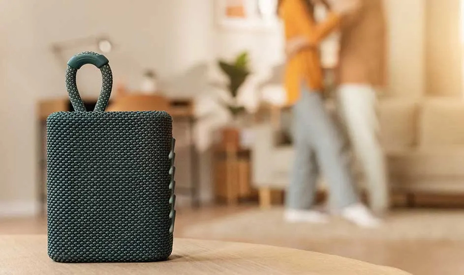 Au premier plan, une enceinte noire compacte et tressée avec une boucle repose, tandis qu’une personne marche dans un intérieur moderne.
