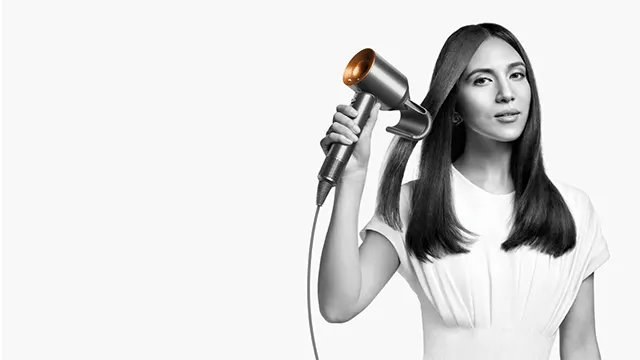 Une femme utilise un sèche-cheveux sur fond blanc. Le sèche-cheveux est argenté avec une buse dorée. Elle porte un t-shirt blanc.