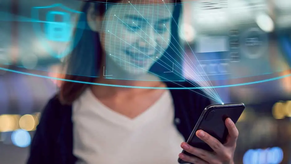 Une personne tient un smartphone noir avec des projections bleues, suggérant une reconnaissance faciale. L'arrière-plan est flou.