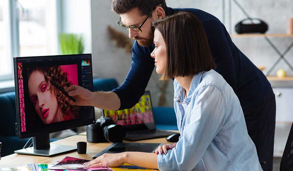 A man and a woman sit at a desk, closely editing a colorful portrait photo together on a large computer screen.