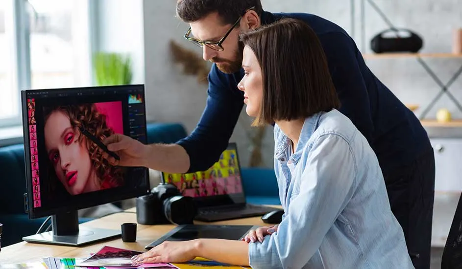 A man and a woman sit at a desk, closely editing a colorful portrait photo together on a large computer screen.