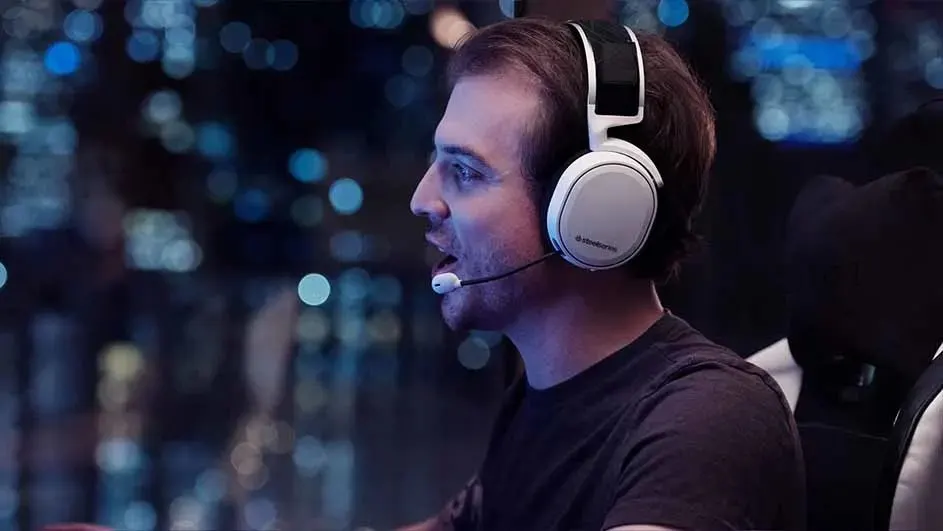 Un homme porte un casque de jeu blanc avec microphone, devant un fond flou de lumières bleues.
