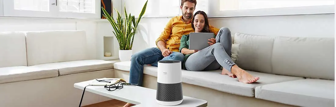 A relaxed couple sits on a bright sofa with a tablet, while a modern air purifier on the coffee table quietly refreshes the living room air.
