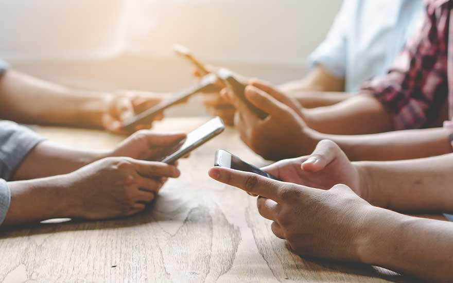 Meerdere handen houden smartphones boven een houten tafel. De achtergrond is vaag en licht.