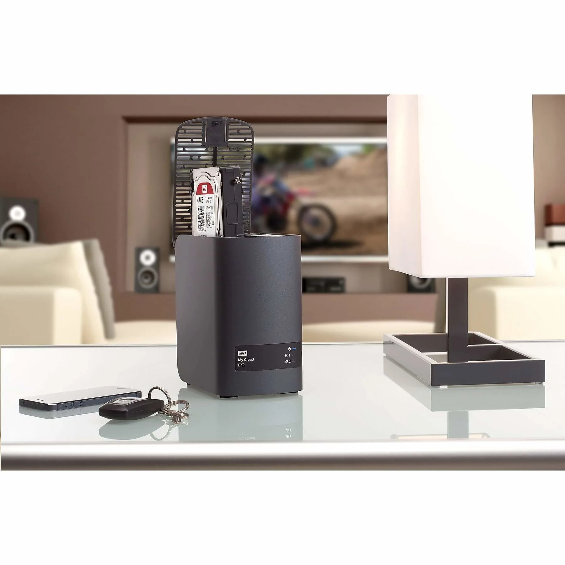 Op een glazen tafel staat een Western Digital My Cloud EX2, een telefoon, sleutels en een lamp. Op de achtergrond staat een tv.