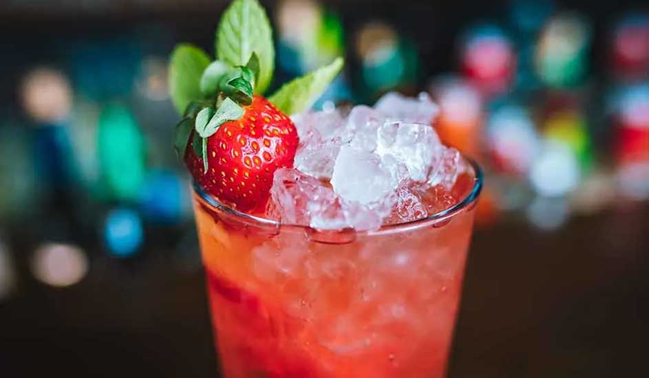 Un verre rempli de glace et d'une boisson rouge, décoré d'une fraise et de feuilles vertes, sur un fond flou coloré.