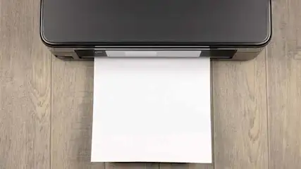 Une imprimante noire imprime une feuille blanche sur une surface en bois clair.