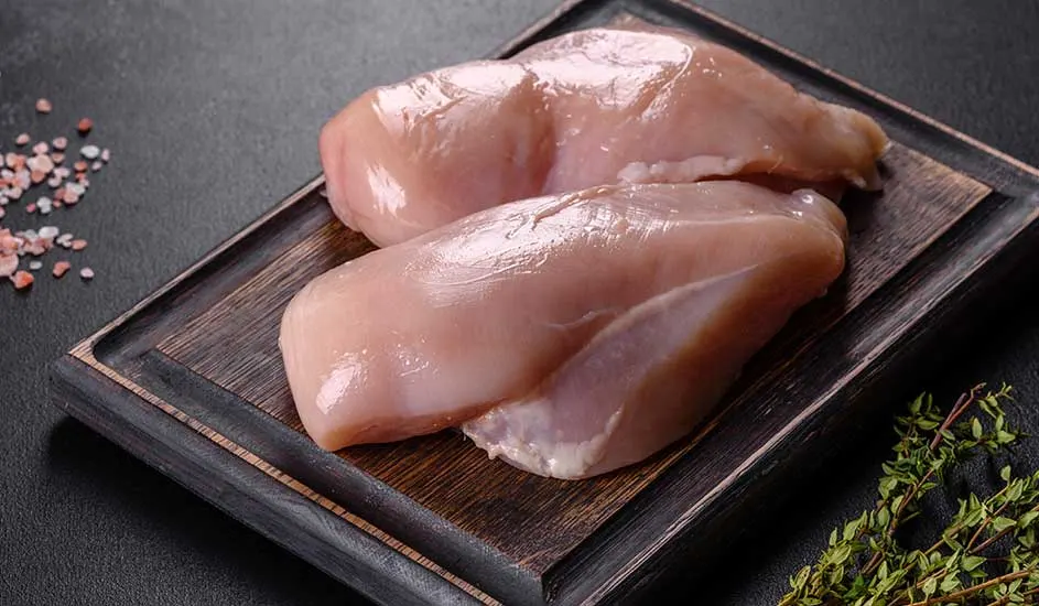Deux filets de poulet crus sur une planche en bois sombre, avec des cristaux de sel rose et du thym.