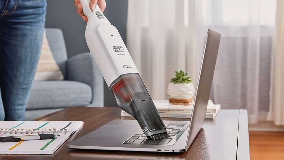 Une personne utilise un aspirateur à main blanc sur un ordinateur portable argenté. Un bloc-notes et un stylo sont sur le bureau.