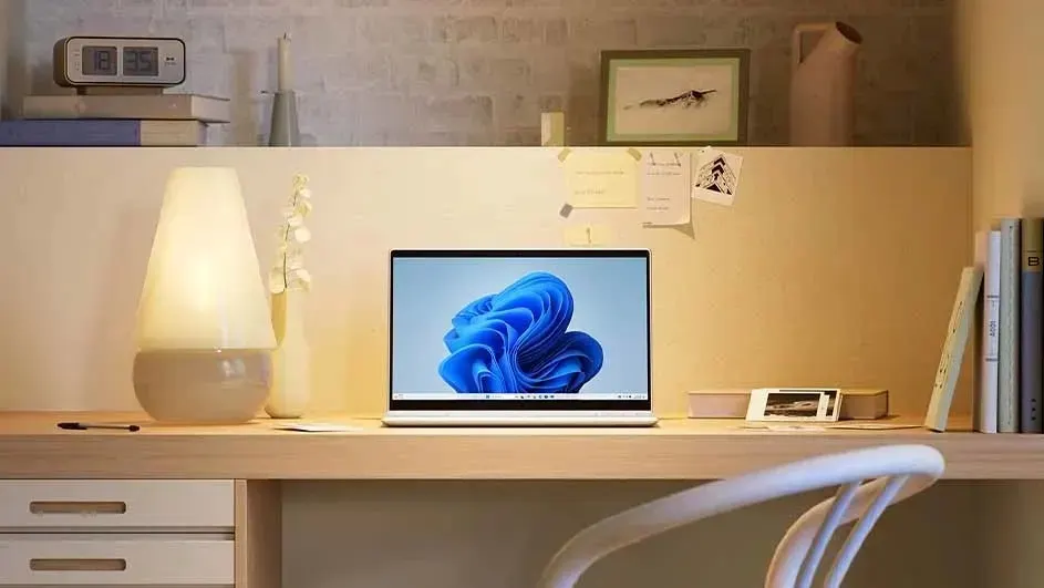 Bright wooden desk with open laptop centered, glowing lamp left, books and photos right, creating a calm, tidy home office.