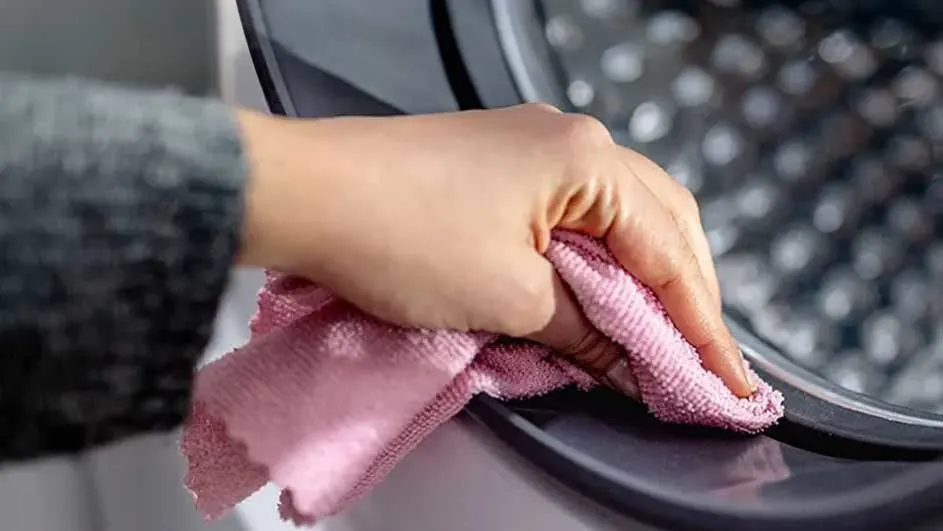 Une main essuie un hublot de machine à laver gris foncé avec un chiffon rose. L'intérieur est visible.