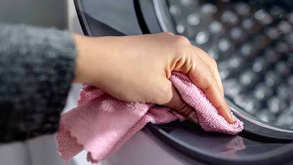 Een hand met een roze doek maakt een grijze wasmachine schoon. De trommel is zichtbaar op de achtergrond.
